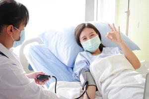 pacientes do sexo feminino asiáticos usam uma máscara azul e deitam em uma cama de hospital. Levante sua mão. OK. o médico mede a pressão do paciente. conceito de serviço médico, seguro de saúde foto