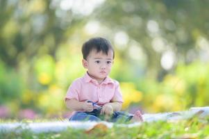 menino asiático sentado no tapete segurando uma caneta enquanto aprende de fora da escola no parque natural foto