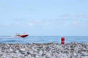 antalya, turquia - 18 de maio de 2021 lata vermelha original da coca cola encontra-se em pequenas pedras redondas perto da costa do mar. lata de coca-cola e lancha na praia foto