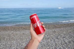 antalya, turquia - 18 de maio de 2021 lata vermelha original da coca cola encontra-se em pequenas pedras redondas perto da costa do mar. lata de coca-cola na mão masculina na praia foto