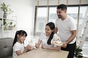 uma família tailandesa asiática saudável, filha e jovem mãe bebem leite branco fresco em vidro e pão do pai, alegria juntos em uma mesa de jantar de manhã, bem-estar nutrição em casa café da manhã refeição estilo de vida. foto
