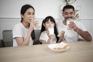 uma família tailandesa asiática saudável, uma filha pequena e pais jovens bebem leite branco fresco em vidro e pão alegria juntos em uma mesa de jantar de manhã, bem-estar nutrição casa café da manhã refeição estilo de vida. foto