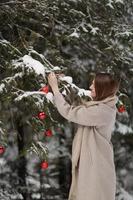 linda garota decora a árvore de Natal com bolas vermelhas na floresta. madeira de inverno. inverno. feliz Natal foto