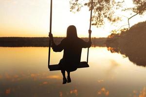 jovem romântica em um balanço sobre o lago ao pôr do sol. jovem viajante sentada no balanço na bela natureza, vista para o lago foto