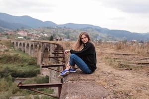 garota jovem turista sentado no viaduto com trilhos antigos. antigo viaduto ferroviário na aldeia estância de montanha de vorokhta. Ucrânia, Cárpatos. foto