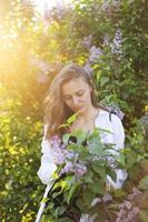 irreal deslumbrante mulher jovem e bonita andando em um verde florido na primavera em um jardim de lavanda com um buquê de lilás nas mãos. closeup retrato romântico de moda. cheirando as flores foto
