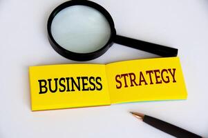 texto de estratégia de negócios no bloco de notas amarelo com lupa e caneta sobre fundo branco. conceito de estratégia de negócios foto