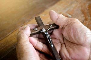 A cruz de madeira closeup que tem uma estátua de metal de jesus crucificado está nas mãos de um católico ancião asiático enquanto reza em uma igreja local, foco suave e seletivo. foto