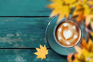 bebendo café na temporada de outono e outono. xícara de café com leite quente na mesa de madeira azul. vista do topo. concentre-se em latte art em forma de coração. folha de bordo amarela turva como primeiro plano foto