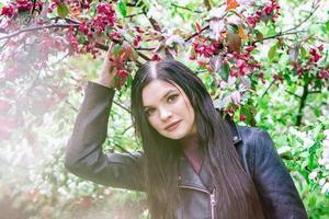 retrato de uma mulher atraente com cabelos castanhos compridos na jaqueta de couro no jardim com flores de macieira flor vermelha e gotas de chuva. foto