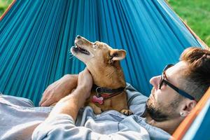 homem europeu bonito em óculos de sol está descansando na rede com seu cachorrinho fofo. foto