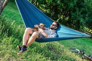 jovem atraente europeu está cochilando na rede com seu cachorrinho fofo. fim de semana ensolarado no parque de verão. foto