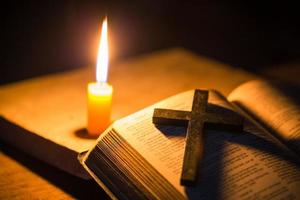 luz de vela com a bíblia sagrada e cruz ou crucifixo sobre fundo de madeira velho na igreja. luz de velas e livro aberto no estudo de cristianismo de mesa de madeira vintage e leitura em casa. conceito de religião de cristo foto