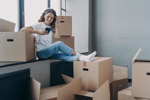 garota sorridente usa aplicativos de telefone compram móveis para nova casa, pesquisa empresa de mudanças. comércio eletrônico, realocação foto