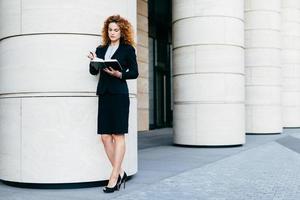 magro jovem empresária em roupas elegantes e sapatos de salto alto, tendo concentrado olhar em seu caderno, encontrando data livre para reunião. freelancer feminino sério com seu diário e caneta foto