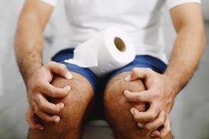 homem sofre de diarréia mão segure o rolo de papel de seda na frente do vaso sanitário. constipação no banheiro. tratamento dor de estômago e higiene, cuidados de saúde foto