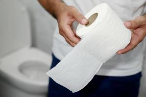 homem sofre de diarréia mão segure o rolo de papel de seda na frente do vaso sanitário. constipação no banheiro. tratamento dor de estômago e higiene, cuidados de saúde foto