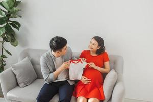 jovem marido e uma esposa grávida mostram sua camiseta de bebê quando estão sentados no sofá. foto