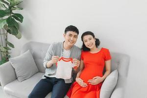 retrato de uma mulher grávida feliz segurando a camiseta do bebê e do marido em um sofá em casa foto