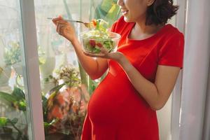 jovem grávida comendo salada de legumes perto da janela em casa foto