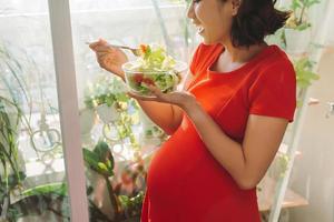 cheio de vitaminas. enérgica mulher grávida bonita comendo sua carne enquanto carregava o prato na mão e relaxando contra a janela foto