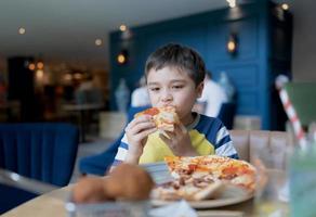 garoto de retrato comendo pizza caseira no café, menino criança feliz mordendo grande fatia de pizza fresca no restaurante, conceito de tempo feliz em família foto