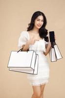 jovem mulher asiática elegante mostrando o celular de tela em branco segurando sacolas de compras isoladas em fundo bege e espaço de cópia. compras de pagamento on-line no smartphone. foto