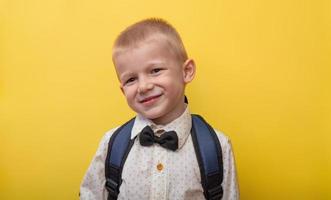 de volta à escola. um menino engraçado loiro com uma mochila em uma camisa leve em um fundo amarelo sorri e olha para a câmera. copie o espaço. foto