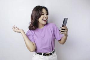 um retrato de uma mulher asiática feliz está sorrindo e segurando seu smartphone vestindo uma camiseta roxa lilás isolada por um fundo branco foto