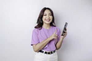 um retrato de uma mulher asiática feliz está sorrindo e segurando seu smartphone vestindo uma camiseta roxa lilás isolada por um fundo branco foto