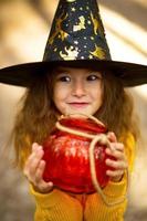 uma garota com um chapéu de bruxa, um suéter amarelo e com uma abóbora uma lanterna e um balde para doces na floresta de outono. feriado de dia das bruxas. retrato, close-up foto