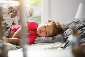 uma criança pequena dorme docemente em sua cama. sono saudável da menina, ventilação. manhã, acordar, luz do sol da janela. ascensão precoce ao jardim de infância, escola. foto