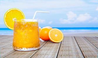 suco de laranja em um copo transparente com cubos de gelo e suco de laranja na boca do copo. bebidas geladas colocadas na varanda de madeira à beira-mar no meio do dia. renderização em 3D foto