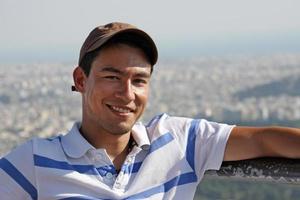 jovem bonito e sorridente sentado ao sol com a cidade de atenas, grécia, ao fundo foto