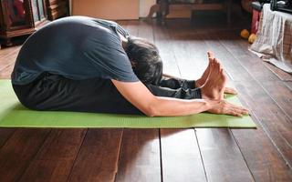 jovem asiático praticando ioga, sentado no exercício de flexão para a frente sentado, pose de paschimottanasana na ioga de esteira verde em casa de madeira. vida saudável foto