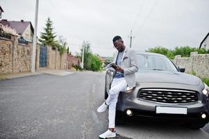 homem afro-americano rico e elegante de blazer e calça branca, óculos lê revista contra seu carro suv. foto