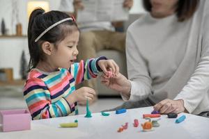 mãe com sua filha brincando com uma argila de modelagem de brinquedos coloridos no chão, no fundo papai está sentado em um sofá na sala de estar, família asiática feliz em casa foto