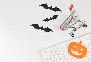 postura plana de carrinho de compras, teclado de computador, decorações de halloween, morcegos e abóbora em fundo branco com espaço de cópia. foto