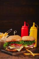 dois hambúrgueres caseiros com marmelada de carne, queijo e cebola em uma placa de madeira, batatas fritas em uma cesta de metal e molhos. conceito de fast food, comida americana foto