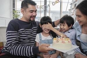 feliz família tailandesa asiática, filha jovem é surpreendida com bolo de aniversário, sopra vela, reza e alegre celebra festa com os pais juntos na sala de estar, estilo de vida de eventos domésticos domésticos de bem-estar. foto