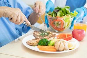 paciente idosa asiática ou idosa tomando café da manhã e comida saudável vegetal com esperança e feliz enquanto está sentado e com fome na cama no hospital. foto