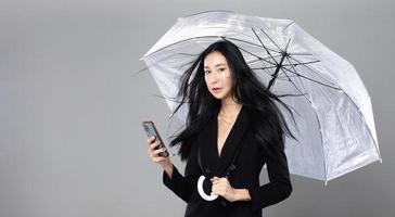 mulher transgênero asiática com longos cabelos lisos pretos, golpe de vento no ar. fêmea segura telefone e guarda-chuva contra tempestade de vento, sentindo moda sensual sexy, espaço de cópia isolado de fundo cinza foto