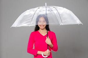 mulher asiática jovem adulta de 20 anos com longos cabelos lisos e pretos usam vestido rosa. fêmea segura guarda-chuva transparente, sentindo emoção moda sorriso feliz, espaço de cópia isolado de fundo cinza foto