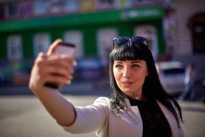 mulher bonita com óculos escuros e cabelos compridos está usando o dispositivo móvel aproveitando os raios de sol e o dia quente ao ar livre. retrato de morena sorridente fazendo selfie em um smartphone foto