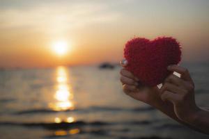 mão segurando o coração de artesanato vermelho no mar no pôr do sol da noite. foto