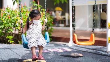 linda garota asiática está jogando no balanço no playground ao ar livre. durante o verão ou a primavera. as crianças usam máscaras para evitar a poluição por poeira, fumaça ou propagação de vírus no ar. crianças de 5 a 6 anos. foto