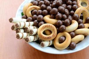 túbulos crocantes, bolas de chocolate derretendo e bagels estão em um prato branco sobre uma mesa de madeira. mistura de vários doces foto