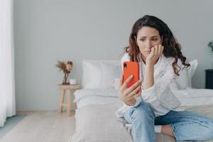 mulher perplexa chateada lendo más notícias da mídia em redes sociais segurando smartphone sentado no quarto foto