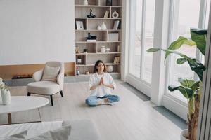 fêmea medita praticando ioga sentada no chão, respirando ar fresco na moderna sala de estar em casa foto