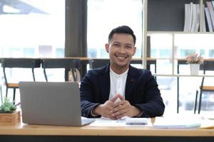 retrato de um jovem empresário asiático sorrindo enquanto estiver usando um laptop e escrevendo notas enquanto está sentado em sua mesa em um escritório moderno